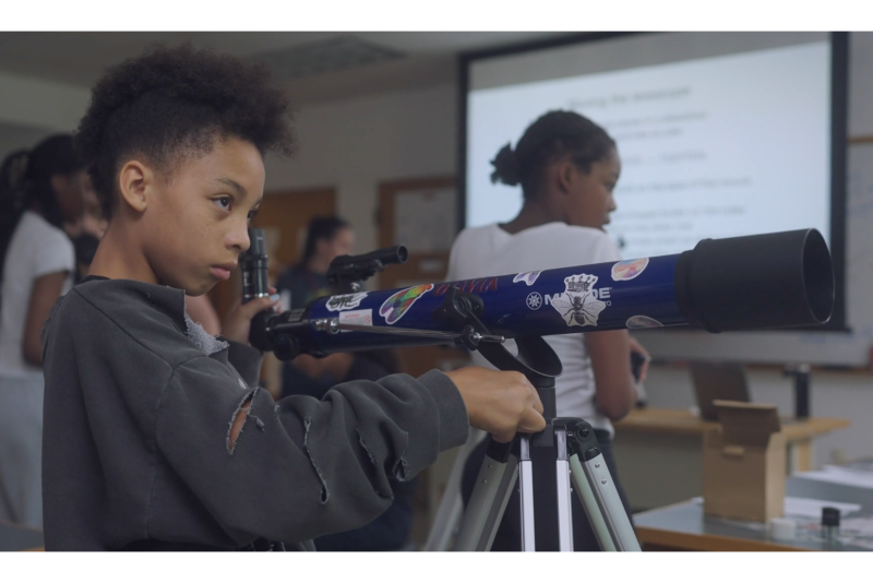 A young girl adjusts a large blue telescope in a classroom.