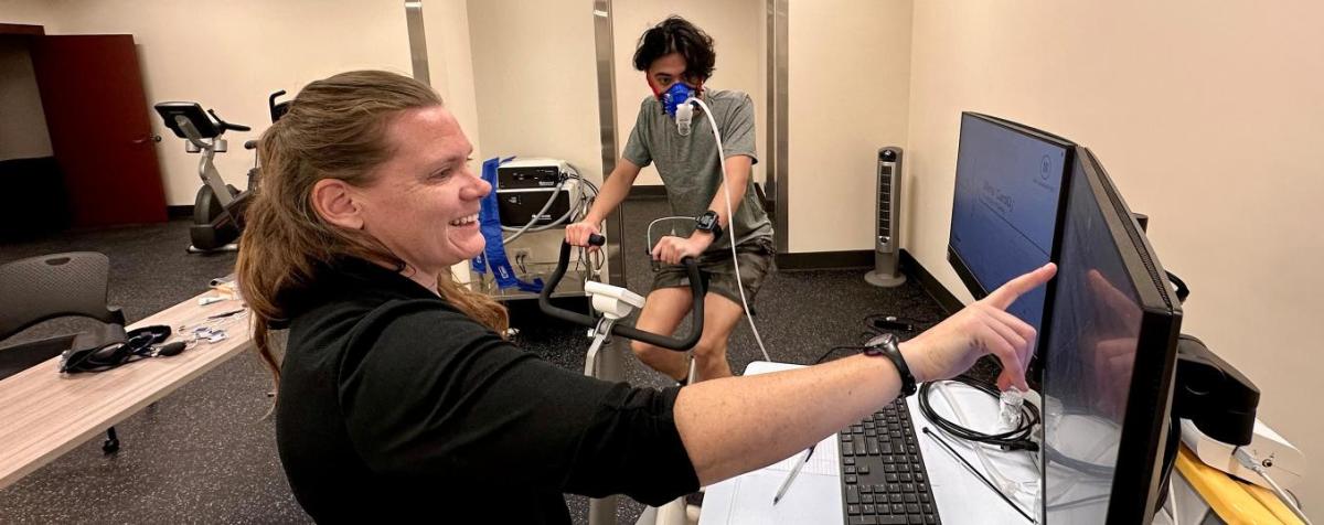 Professor pointing at screen with man on exercise bike in background