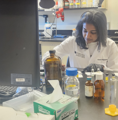 A graduate researcher in Kumar's lab in a lab coat with glass bottles on a lab table in front of her.