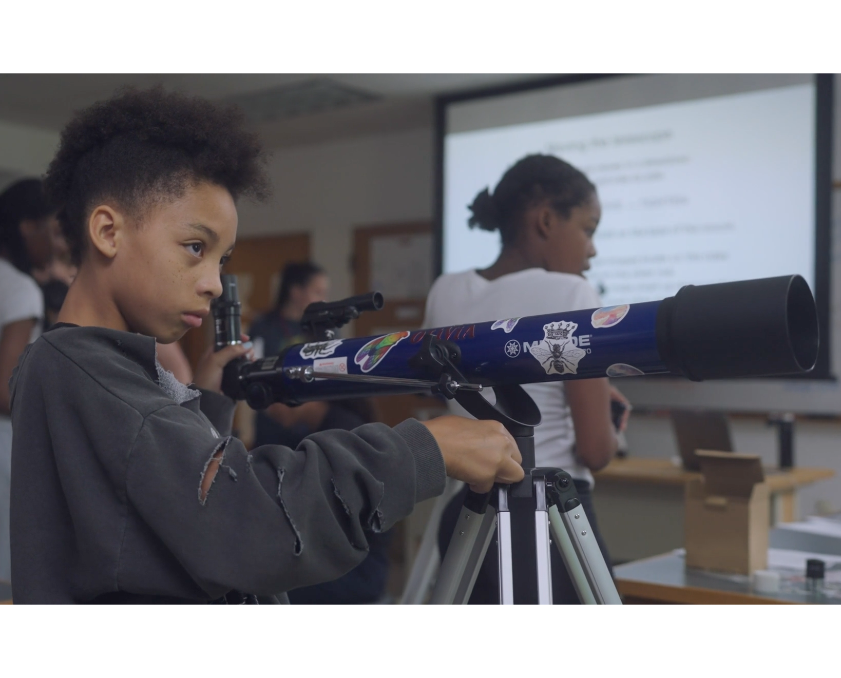 A young girl adjusts a large blue telescope in a classroom.